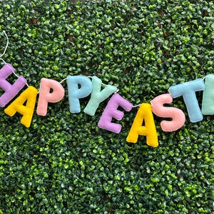 May include: A felt banner with the words "Happy Easter" in pastel colors. The letters are strung together with white thread and are hanging against a green background.