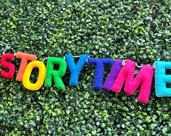 Puede incluir: Una colorida pancarta de fieltro que deletrea "STORYTIME" en tonos arcoíris sobre un fondo de follaje verde. Las letras están cortadas y cosidas individualmente, creando un acento alegre y decorativo para un espacio o evento infantil.