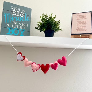 K&ouml;nnte beinhalten: Eine wei&szlig;e Schnur mit einer Girlande aus rosa und roten Filzherzen, die an einem wei&szlig;en Regal h&auml;ngt. Das Regal hat ein graues Schild mit dem Text "Such a Big Miracle in Such a Little Boy" und einen kleinen gerahmten Druck mit spanischem Text.