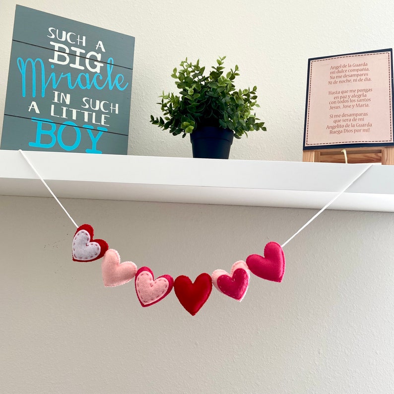 K&ouml;nnte beinhalten: Eine wei&szlig;e Schnur mit einer Girlande aus rosa und roten Filzherzen, die an einem wei&szlig;en Regal h&auml;ngt. Das Regal hat ein graues Schild mit dem Text "Such a Big Miracle in Such a Little Boy" und einen kleinen gerahmten Druck mit spanischem Text.