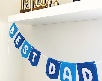 Könnte beinhalten: Eine blaue Wimpelkette mit den weißen Buchstaben "BEST DAD". Die Kette ist aus Filz und an einer weißen Schnur befestigt. Ein Holzschild mit der Aufschrift "Faith" befindet sich auf einem Regal darüber.
