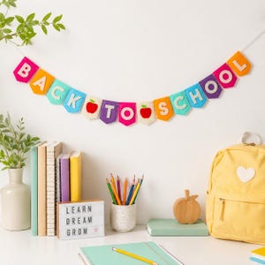 Peut inclure: Une bannière en feutrine colorée affichant le texte BACK TO SCHOOL avec deux icônes de pommes rouges, suspendue à une ficelle. Les lettres sont de diverses couleurs vives dont rose, jaune, bleu clair, violet et orange, en forme de fanions.