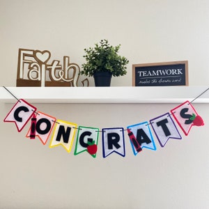 Puede incluir: Una pancarta de fieltro de colores con la palabra "CONGRATS" en letras negras. Cada letra está decorada con una manzana roja o un crayón rojo. La pancarta está colgada de una repisa blanca con un letrero de madera que dice "Faith" y una pequeña planta en maceta.