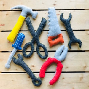 May include: A set of felt toy tools, including a hammer, scissors, pliers, a screwdriver, and a saw. The tools are all different colours and have a playful, whimsical design.