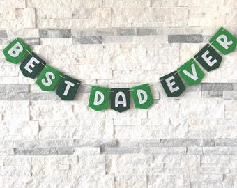 Könnte beinhalten: Grüne Wimpelkette mit den weißen Buchstaben "BEST DAD EVER". Die Wimpel sind an einer weißen Schnur befestigt und hängen vor einer weißen Backsteinmauer, ideal für Vatertag.
