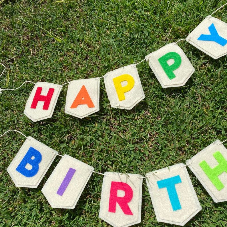 Pu&ograve; includere: Un festoso striscione con la scritta "HAPPY BIRTHDAY" in lettere di feltro colorate. Le lettere sono attaccate a piccoli stendardi bianchi sporchi collegati da uno spago bianco, su uno sfondo di erba verde.