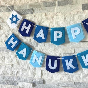 Puede incluir: Una guirnalda festiva de Janucá con banderines de fieltro azul y blanco que deletrean "HAPPY HANUKKAH". La pancarta incluye una Estrella de David y está ensartada en un cordón blanco. Los banderines son de varios tonos de azul.