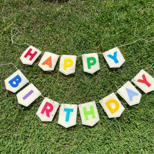 Pu&ograve; includere: Uno striscione festivo "Buon Compleanno" con lettere colorate su singoli pezzi a forma di bandiera. Le lettere sono rosse, arancioni, gialle, verdi, blu, viola e azzurre. Lo striscione &egrave; realizzato in tessuto beige chiaro ed &egrave; infilato su una corda bianca.