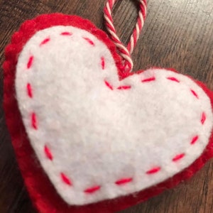 Op de afbeelding: Een rood vilt hart met een wit vilt middenstuk en roze stiksels rond de randen. Het hart heeft een wit en rood touwtje dat aan de bovenkant is bevestigd om het op te hangen.