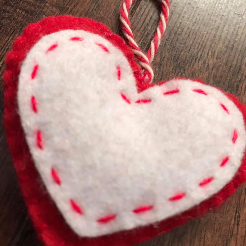 Op de afbeelding: Een rood vilt hart met een wit vilt middenstuk en roze stiksels rond de randen. Het hart heeft een wit en rood touwtje dat aan de bovenkant is bevestigd om het op te hangen.