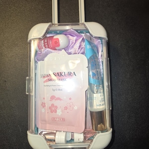 May include: A clear, miniature suitcase filled with beauty products. The suitcase has a white handle and wheels. Inside are pink and white packets of Japan Sakura Mud Mask, a lip balm, and other skincare items.