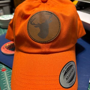 May include: An orange baseball cap with a brown leather patch featuring a deer silhouette. The patch is stitched onto the front of the cap. The cap is adjustable and has a black label that says "The Authentic Yupoong Adjustable".