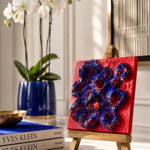 Peut inclure: Petite peinture abstraite rouge avec des détails texturés bleus et rouges, présentée sur un chevalet en bois miniature. La peinture est placée devant un mur blanc, un vase bleu avec des orchidées blanches et des livres.
