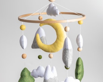 May include: A wooden baby mobile with felt clouds, mountains, trees, and a crescent moon. The mobile is suspended from a white metal arm.