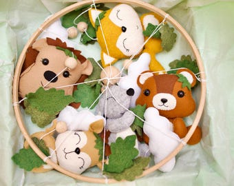May include: A wooden hoop mobile with felt woodland animal characters. The mobile features a fox, a squirrel, a bear, a hedgehog, and a koala. The animals are all sewn with felt and have embroidered details. The mobile is hung from a wooden hoop and has green felt leaves.