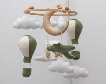 Puede incluir: Un globo aerostático verde y blanco, un avión verde, una luna creciente dorada y nubes blancas cuelgan de un anillo de madera. Este móvil está hecho de fieltro y es perfecto para la habitación de un bebé.