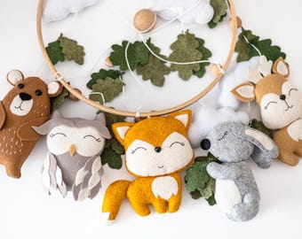 May include: A wooden baby mobile with felt woodland animal characters, including a bear, owl, fox, bunny, and deer. The animals are all smiling and have closed eyes. The mobile is decorated with green felt leaves.