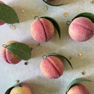 May include: A collection of peach-colored ornaments with green leaves. The ornaments are covered in a sparkly, textured finish and have a delicate, vintage aesthetic.