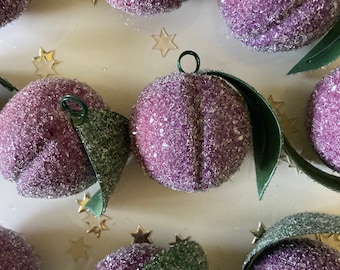 May include: Close-up of several plum-shaped ornaments. Each ornament is a vibrant purple color and covered in glitter, with a green leaf and a metal loop. Gold star-shaped confetti is scattered around the ornaments.