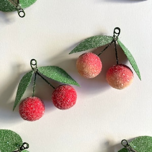 May include: Close-up of several artificial cherries with red and yellow-orange fruit, green leaves, and black wire stems. The cherries have a textured, frosted appearance. The leaves are also textured and have a dusting of white. The background is plain white.