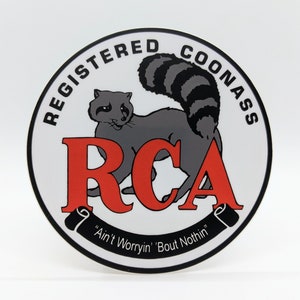 May include: A white circle sticker with black text that reads "Registered Coonass" and "Ain't Worryin' Bout Nothin'" around the edge. A cartoon raccoon with a gray body and black stripes on its tail is in the center of the sticker. The words "RCA" are in red letters in the center of the sticker.