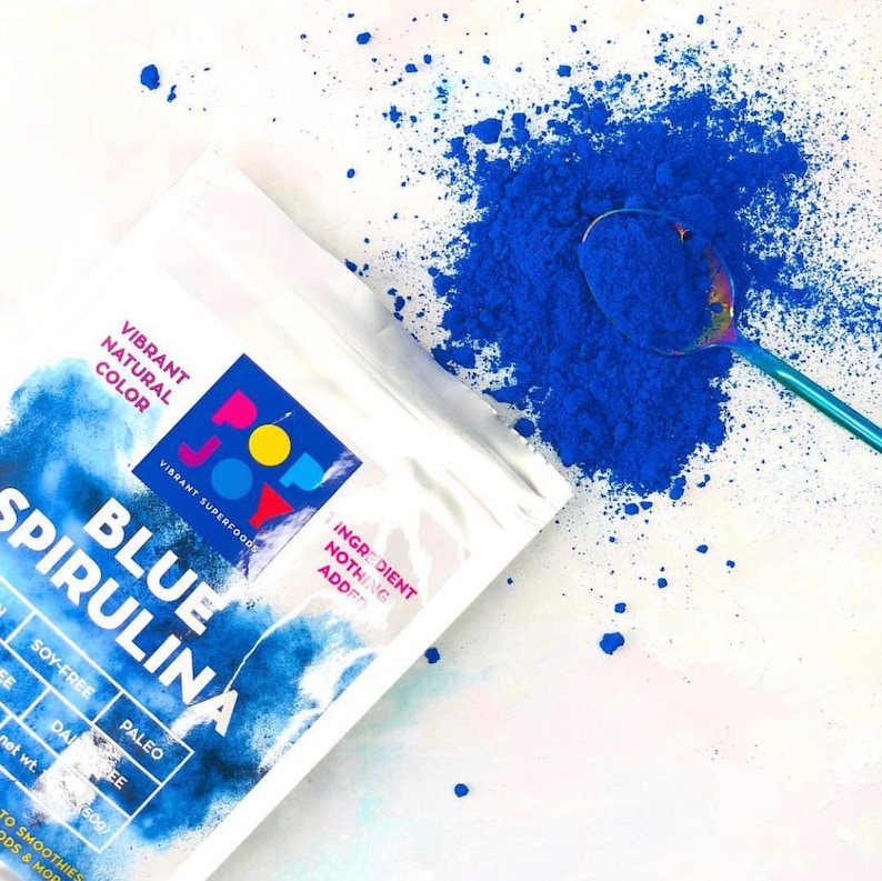 Op de afbeelding: Een witte zak met blauwe tekst die "Pop Joy Vibrant Superfoods Blue Spirulina" en "Ingredient Nothing Added" zegt. De zak is open en blauw poeder valt eruit op een wit oppervlak. Een blauwe lepel ligt op het poeder.