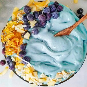 Op de afbeelding: Een blauwe smoothie bowl bedekt met blauwe bessen, ananas en kokosvlokken. De kom is gemaakt van donkerbruin keramiek en heeft een houten lepel die op de rand rust.