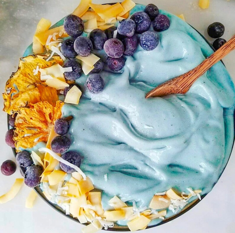 Op de afbeelding: Een blauwe smoothie bowl bedekt met blauwe bessen, ananas en kokosvlokken. De kom is gemaakt van donkerbruin keramiek en heeft een houten lepel die op de rand rust.