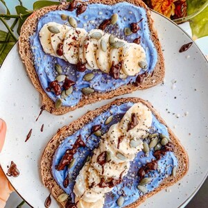 Op de afbeelding: Twee toastjes besmeerd met een paarse roomkaas, gesneden bananen, chocolade chips en pompoenpitten.