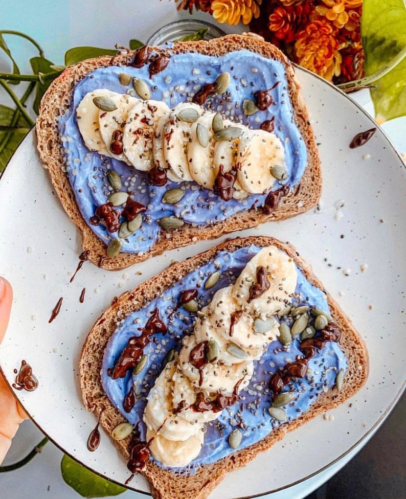 Op de afbeelding: Twee toastjes besmeerd met een paarse roomkaas, gesneden bananen, chocolade chips en pompoenpitten.