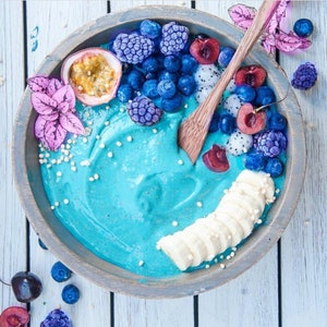 Op de afbeelding: Een blauwe smoothiebowl bedekt met gesneden banaan, blauwe bessen, bramen, kersen en passievrucht. De kom is van hout en heeft een lichtbruine kleur.