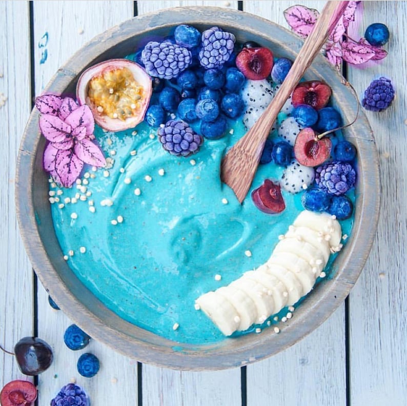Op de afbeelding: Een blauwe smoothiebowl bedekt met gesneden banaan, blauwe bessen, bramen, kersen en passievrucht. De kom is van hout en heeft een lichtbruine kleur.
