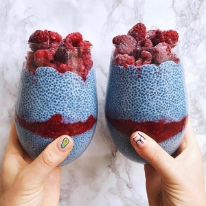 Op de afbeelding: Twee glazen bekers gevuld met blauwe chia-pudding, bedekt met verse frambozen. De chia-pudding is lichtblauw en heeft een gespikkelde textuur.