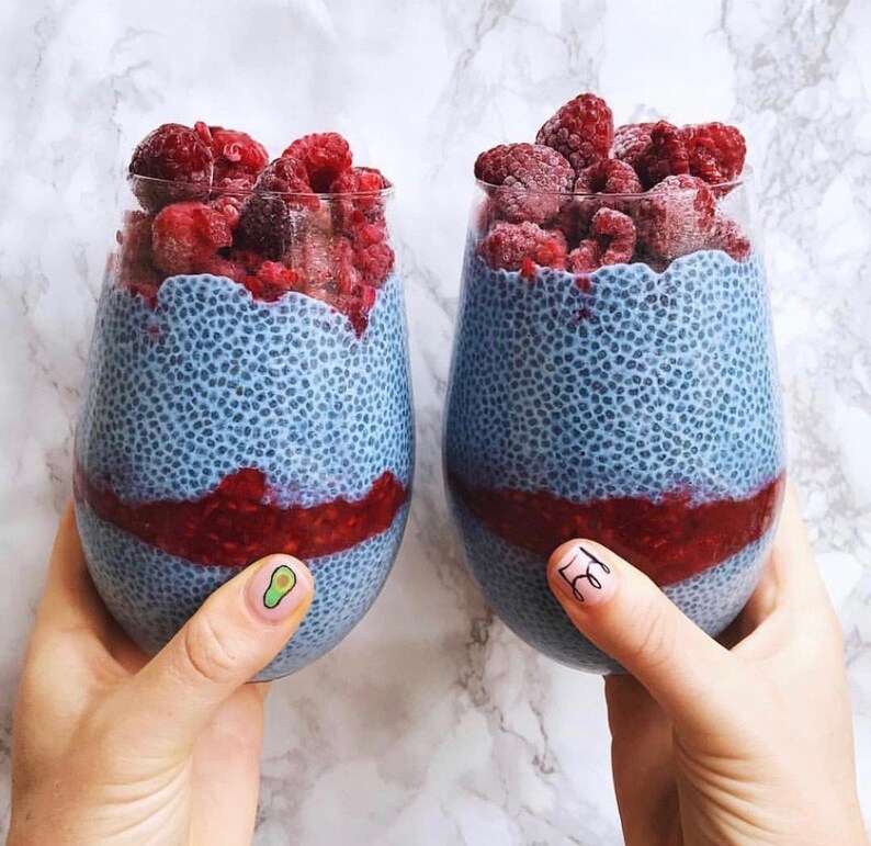 Op de afbeelding: Twee glazen bekers gevuld met blauwe chia-pudding, bedekt met verse frambozen. De chia-pudding is lichtblauw en heeft een gespikkelde textuur.