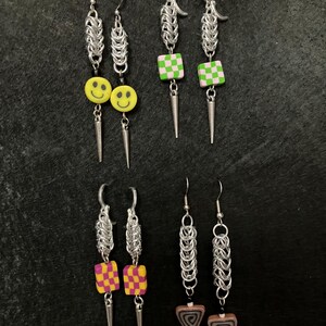 Puede incluir: Una colección de pendientes hechos a mano con varios diseños. Algunos pendientes presentan caras sonrientes, cuadrados a cuadros o formas triangulares con patrones en espiral. Todos los pendientes tienen cota de malla plateada y detalles de púas.