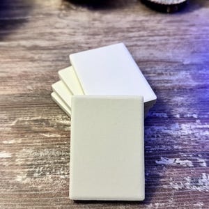 May include: A stack of four rectangular, solid white tiles with smooth, matte surfaces. The tiles are arranged in a slightly fanned-out formation, with the front-most tile positioned vertically and the others layered behind it in a uniform, clean alignment.
