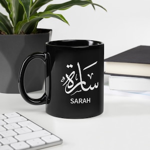 Könnte beinhalten: Schwarze Keramiktasse mit einem weißen arabischen Kalligraphie-Design, das darunter auf Englisch "Sarah" lautet.