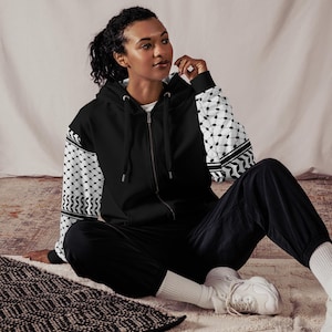 May include: Black zip-up hoodie with patterned sleeves. The sleeves have a black and white geometric design. The person is wearing black trousers and white trainers. The hoodie has a drawstring hood and a full-length zip.