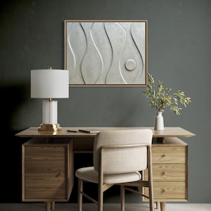 May include: A modern office setup featuring a wooden desk with drawers, a beige chair, and a white lamp with a gold base. A framed abstract wall art piece hangs above the desk. A vase with greenery sits on the desk.