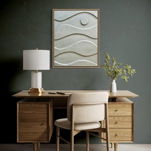 May include: A modern office setup featuring a wooden desk with drawers, a matching chair, and a white lamp with a gold base. A framed abstract wall art with wave-like patterns hangs above the desk. A small vase with greenery adds a touch of nature.