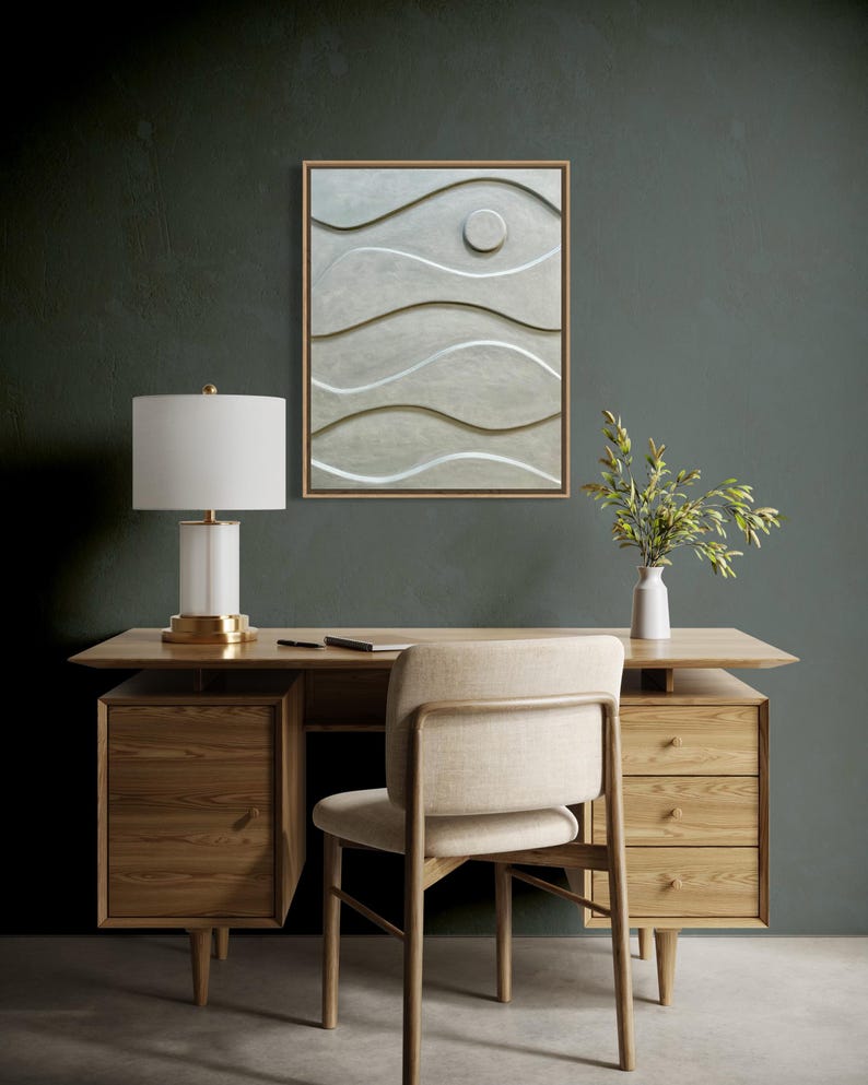 May include: A modern office setup featuring a wooden desk with drawers, a matching chair, and a white lamp with a gold base. A framed abstract wall art with wave-like patterns hangs above the desk. A small vase with greenery adds a touch of nature.