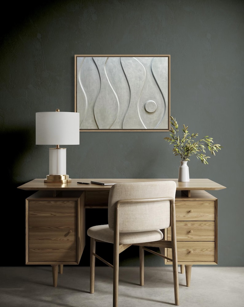 May include: A modern office setup featuring a wooden desk with drawers, a beige chair, and a white lamp with a gold base. A framed abstract wall art piece hangs above the desk. A vase with greenery sits on the desk.
