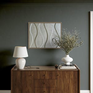 May include: A mid-century modern style wooden sideboard with a white ceramic lamp, a white vase with dried flowers, and a framed abstract art piece. The wall is painted a dark teal color.