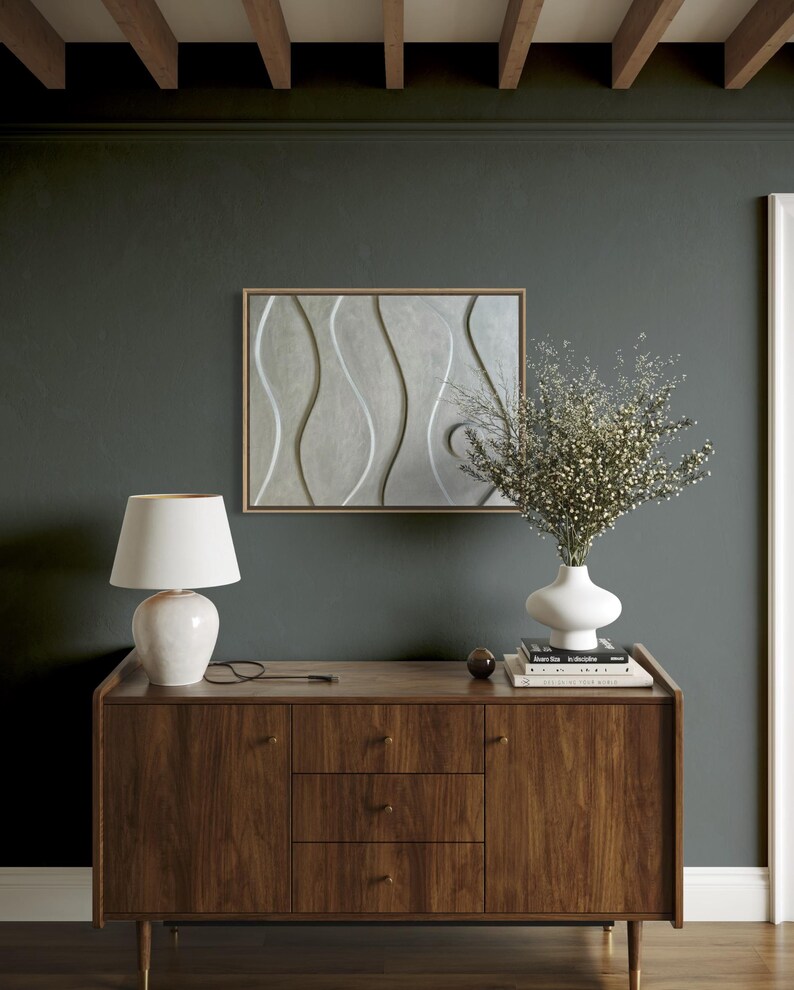 May include: A mid-century modern style wooden sideboard with a white ceramic lamp, a white vase with dried flowers, and a framed abstract art piece. The wall is painted a dark teal color.
