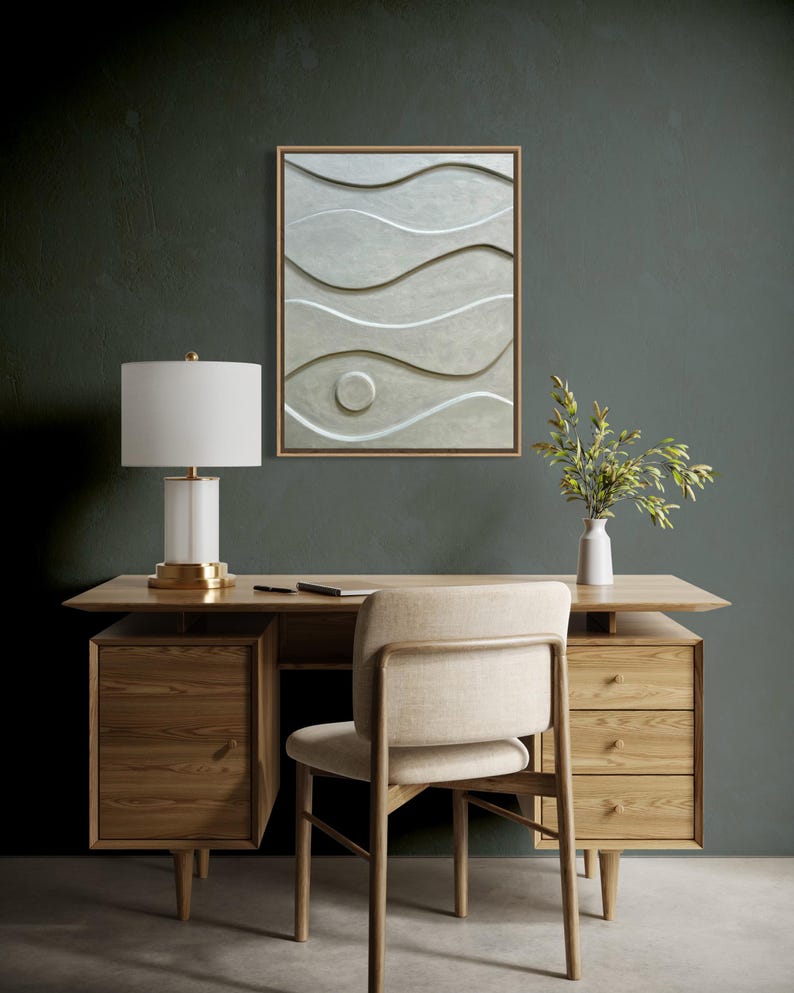 May include: A wooden desk with drawers, a chair, and a white lamp with a gold base. A framed abstract art piece with wave-like patterns hangs on the wall. A small white vase with greenery sits on the desk.