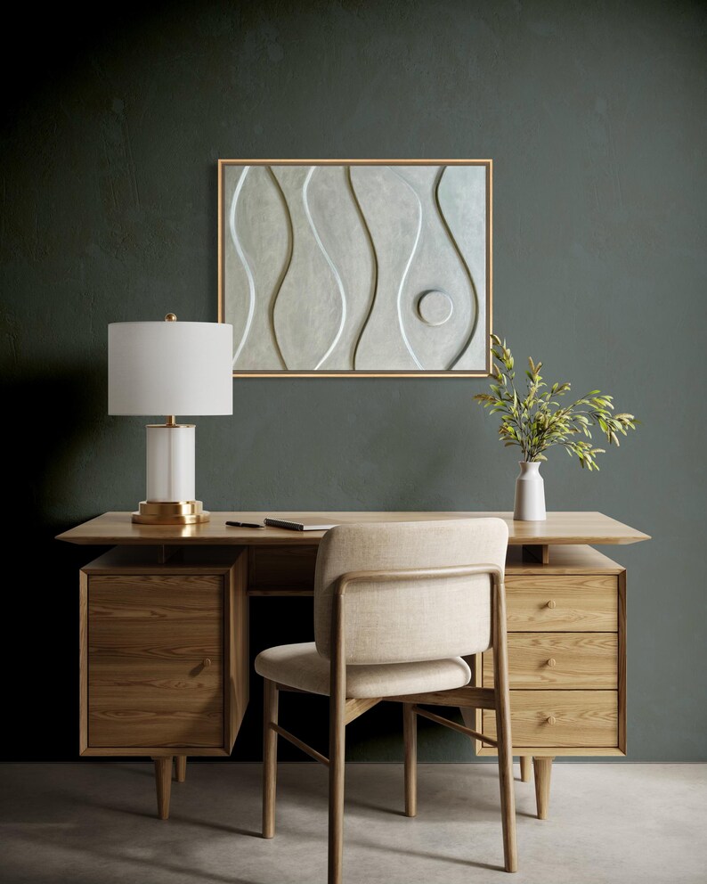 May include: A modern office setup featuring a wooden desk with drawers, a beige chair, and a white lamp with a gold base. A framed abstract wall art with a wavy design hangs above the desk. A vase with greenery sits on the desk.