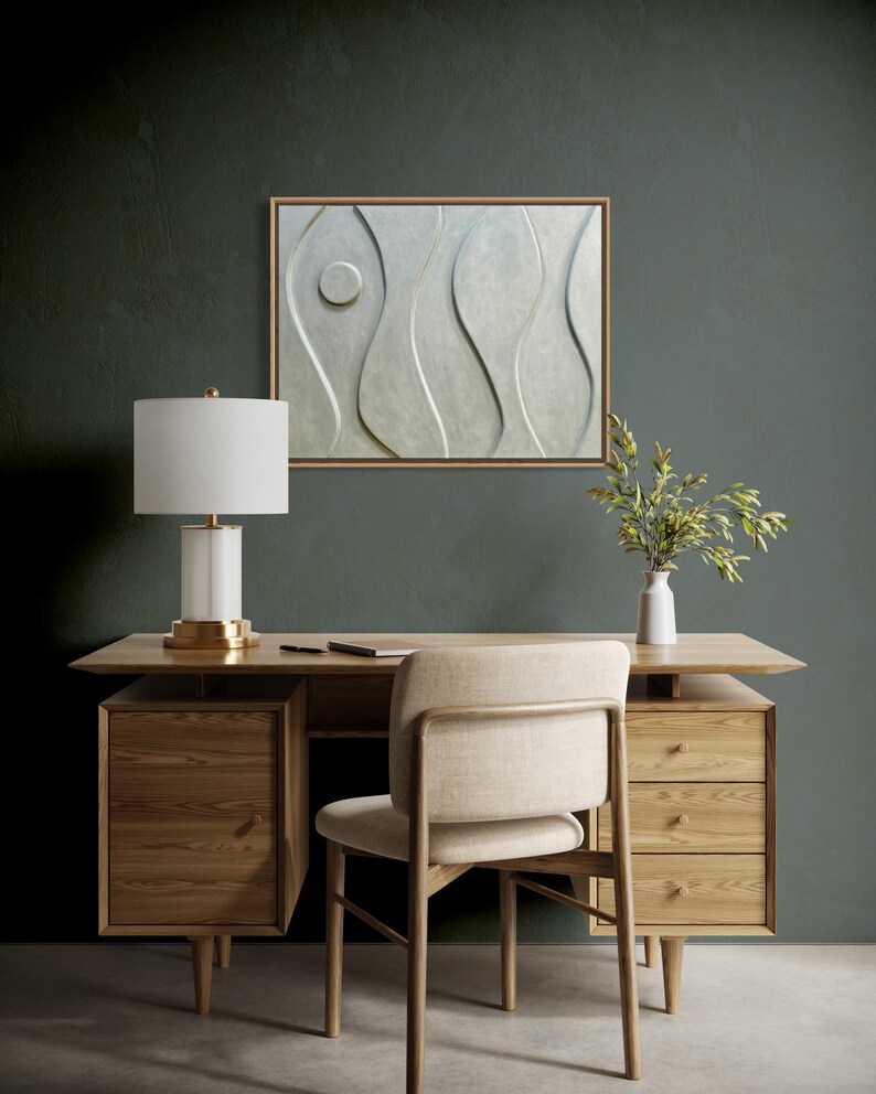 May include: A modern office setup featuring a light wood desk with drawers, a beige chair, and a white lamp with a gold base. A framed abstract wall art piece with a light gray design hangs above the desk. A small white vase with greenery is on the desk.
