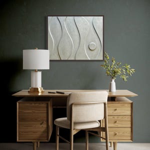 May include: A modern office setup featuring a light wood desk with drawers, a beige chair, and a white lamp with a gold base. A framed abstract wall art piece hangs above the desk. A small white vase with greenery sits on the desk.
