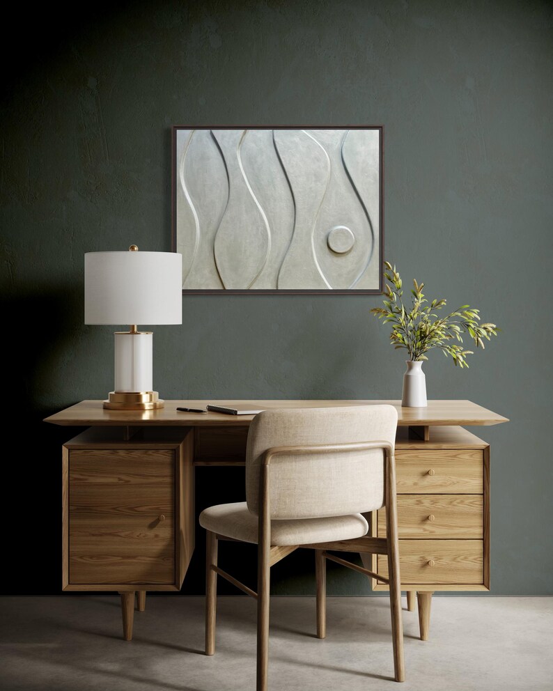 May include: A modern office setup featuring a light wood desk with drawers, a beige chair, and a white lamp with a gold base. A framed abstract wall art piece hangs above the desk. A small white vase with greenery sits on the desk.