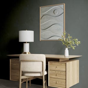 May include: A modern office setup featuring a light wood desk, chair, and a three-drawer cabinet. A white lamp with a gold base sits on the desk. A framed gray artwork with wave-like patterns hangs on the wall. A white vase holds green stems.